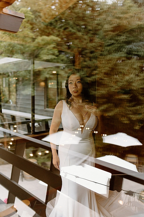 Bridal portrait of a bride in minimalist gown with a deep V neckline and pendant necklace by a window, string lights behind