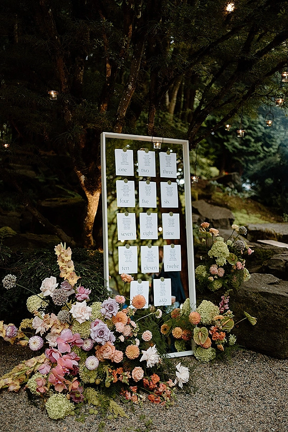 Seating chart mirror seating chart with clipped cards above lush hydrangeas, roses, and dahlias, under string lights by stone steps in a garden
