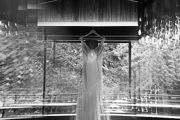 Wedding dress hanging by large windows with a chandelier accent, soft trees outside, showcasing a sleek minimalist bridal gown silhouette