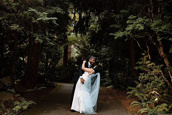 Wedding couple portrait of bride and groom dip kiss on a forest pathway, her long veil flowing beside his black suit and boutonniere