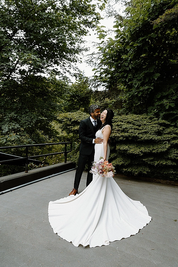 Couple portrait of newlyweds laughing in an embrace, bride holding bouquet with dress train beside a terrace railing and greenery under overcast sky