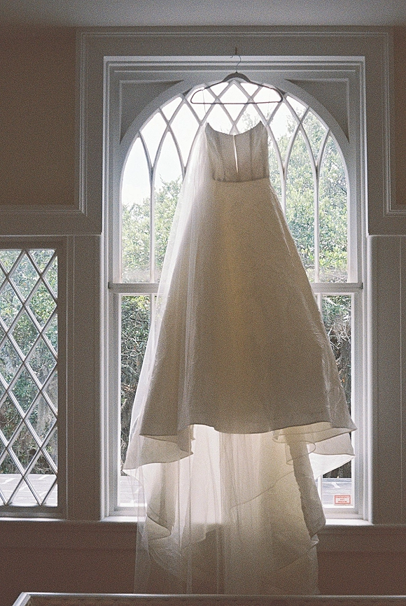 Wedding dress hanging on a dress hanger, a strapless tulle skirted gown lit by natural light near an arched window with trees beyond