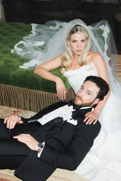 Couple portrait of bride and groom seated pose on a green velvet sofa, her lace-trim veil draped as he lounges in a tuxedo