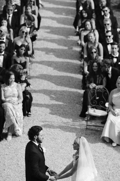 Wedding vows as bride in veil and gown holds hands with groom in tuxedo, guests seated along a gravel aisle outdoors