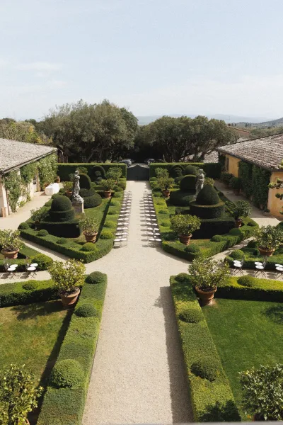 Outdoor ceremony setup with white chairs in neat rows lining a gravel aisle, framed by manicured hedges and stone statues in a formal villa garden