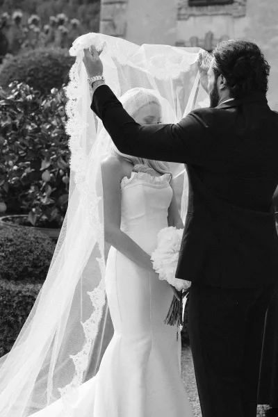 First look moment as groom lifting veil over bride in strapless dress, lace-trim veil and bouquet, in a courtyard by shrubs and stone wall