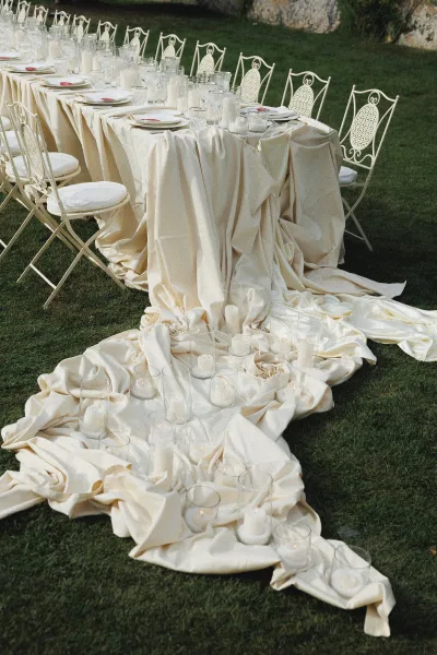 Reception tablescape with draped ivory tablecloth, white plates and clear glassware, red napkin accents, and candles on a lawn by a stone wall