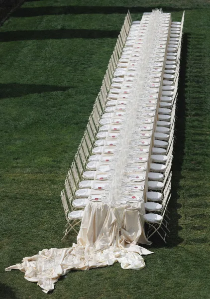 Reception tablescape with long banquet table wedding setup, ivory tablecloth, plates and glassware on a green lawn with white folding chairs