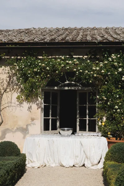 Drink station setup with wedding drink station glassware and a dispenser bowl on a white tablecloth, framed by vines and a lemon tree in a villa courtyard