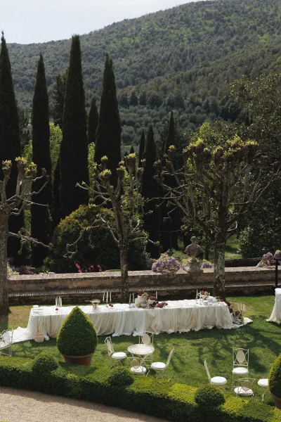 Outdoor reception setup with a long banquet table, white linens, taper candles and florals on a manicured lawn beside a stone terrace wall