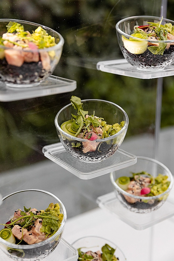 Wedding cocktail appetizers in glass bowls on a clear acrylic riser, bite-size cups with greens and black sesame, set against blurred outdoor foliage