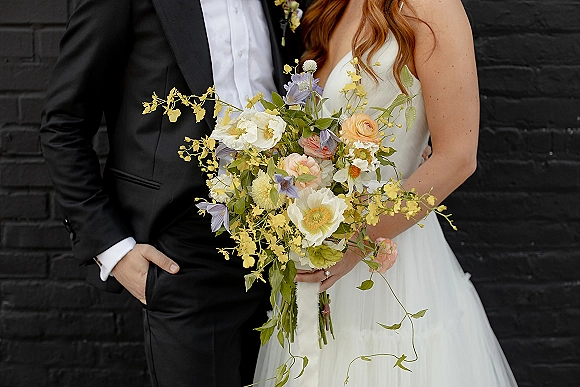 Couple portrait with bride holding bouquet in a strapless wedding dress beside groom in black tuxedo against a black brick wall