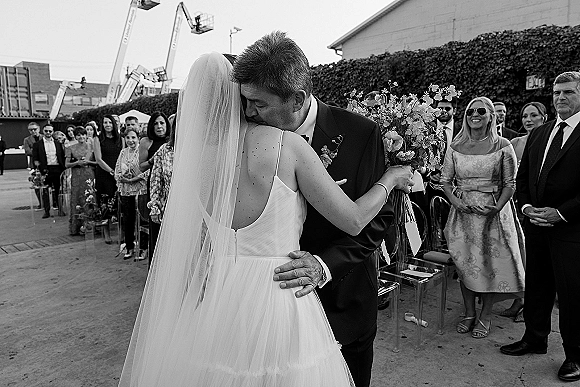 Wedding kiss as bride in veil holds bouquet, groom in tuxedo embraces her in ivy-walled courtyard, guests seated on clear chairs behind