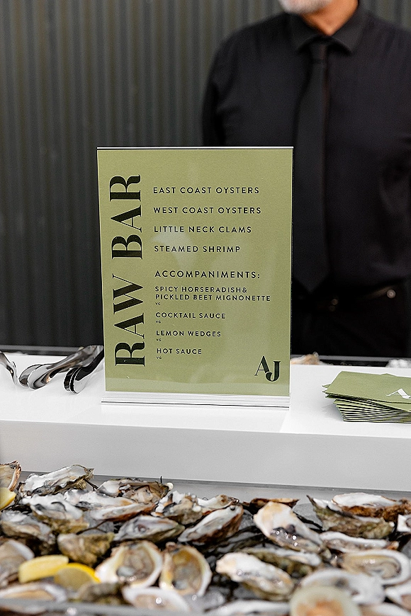 Raw bar sign for wedding raw bar in an acrylic holder beside shucked oysters on ice with lemon wedges, sauces, and tongs on a white counter