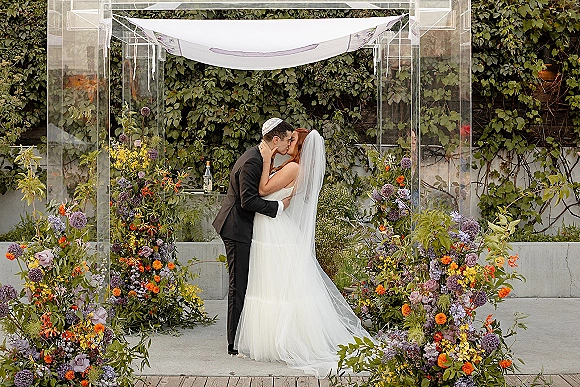 Wedding kiss as bride and groom kiss under a clear acrylic chuppah with white canopy, kippah, veil, and ivy wall backdrop