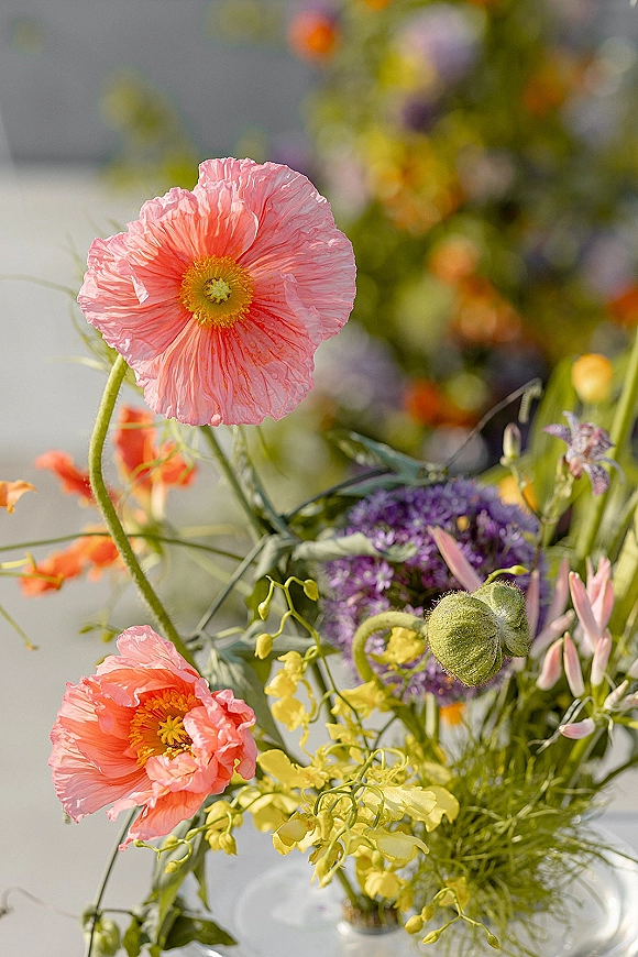 Wedding flowers in a glass vase featuring pink poppy accents with yellow orchid stems and purple allium against a blurred garden backdrop