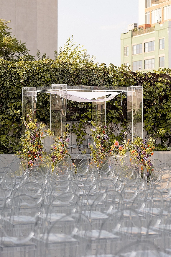 Ceremony setup with an acrylic wedding arch and draped fabric, colorful florals, and clear chairs on a rooftop terrace by an ivy wall