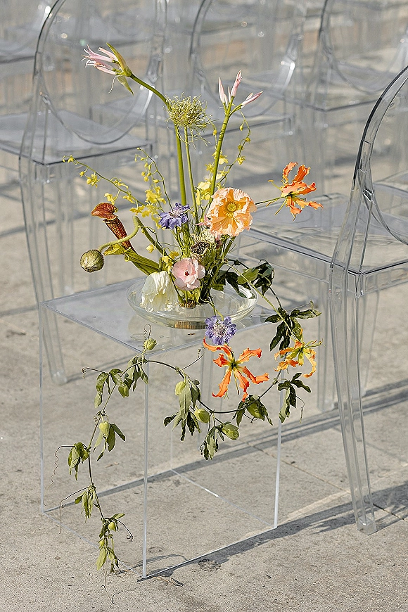 Ceremony chair flowers on a clear acrylic chair with a glass vase, asymmetrical blooms and trailing greenery in outdoor stone patio seating rows