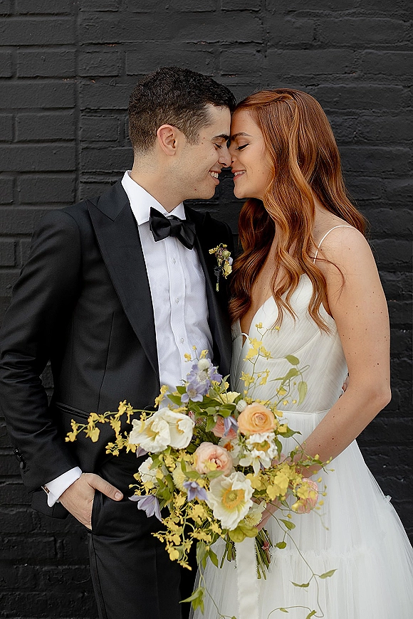 Couple portrait of bride and groom in tuxedo and bridal gown sharing a close embrace with bouquet against a black brick wall backdrop
