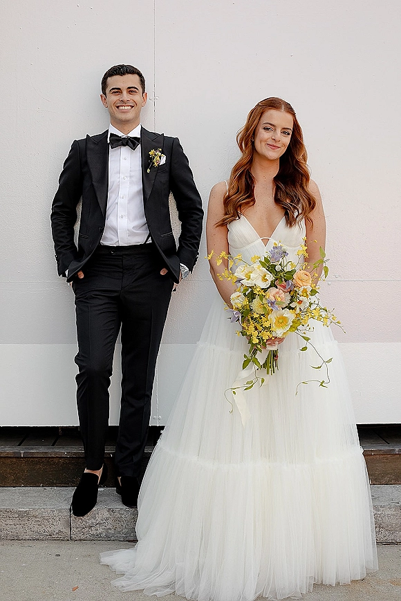 Couple portrait of bride holding bouquet beside groom in black tuxedo, standing on concrete steps against a clean white wall backdrop