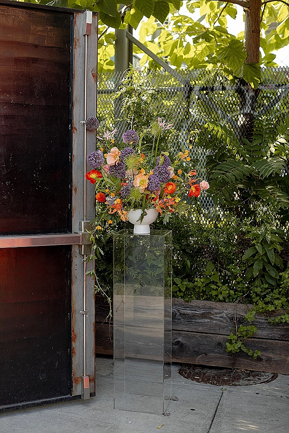 Wedding floral arrangement in a white ceramic bowl on a clear acrylic pedestal, bursting with colorful wildflowers on a concrete patio backdrop