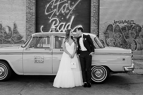 Couple portrait of bride in strapless tulle gown and groom in tuxedo beside a vintage taxi, posed against a graffiti brick wall backdrop