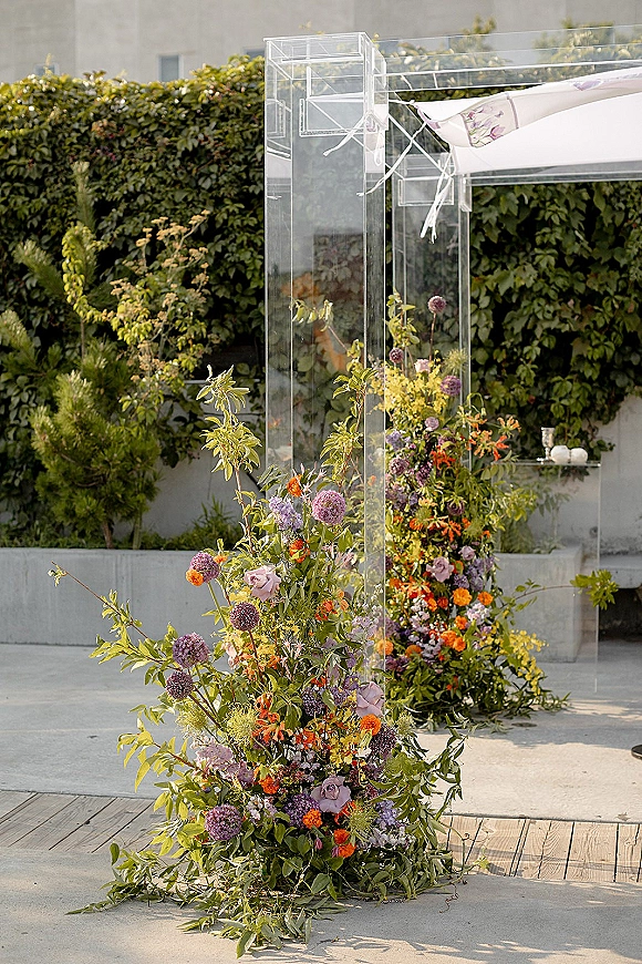 Wedding ceremony backdrop with clear acrylic wedding arch, lush wildflower florals and greenery, set on an outdoor courtyard patio by a hedge