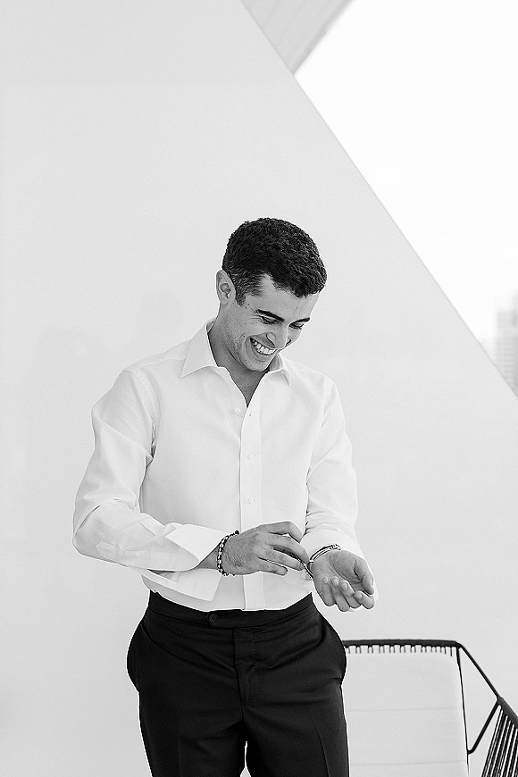 Groom getting ready, adjusting watch over a white dress shirt with bracelet and belt, standing by a chair against a white wall