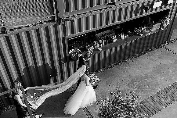 Bride walking in wedding processional moment with bouquet and long veil, groom in suit beside her on an outdoor patio walkway near a bar setup