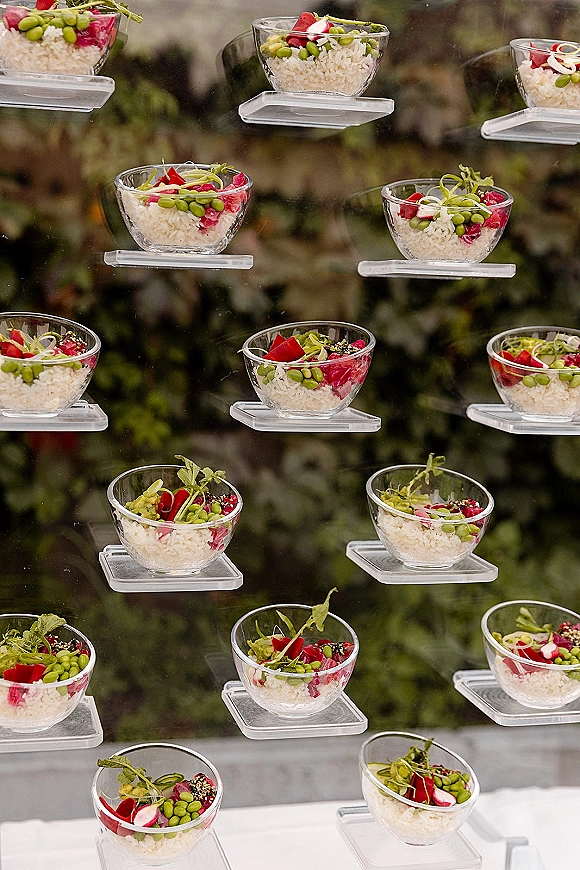 Wedding appetizer display of cocktail hour appetizers in clear cups on acrylic risers, with microgreens and radish, set against garden greenery