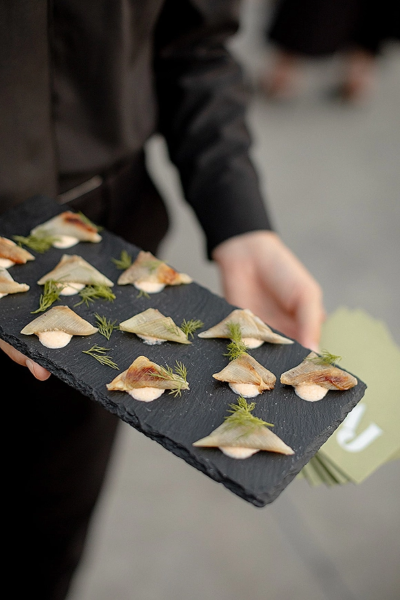 Wedding passed appetizers on a slate serving board with herb garnish and sauce dollops, held by a server on an outdoor patio with greenery