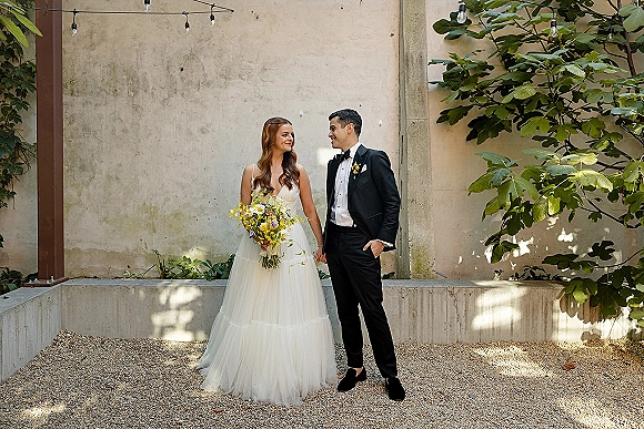 Couple portrait of bride and groom holding hands, she holds a yellow wildflower bouquet, standing by a stucco wall with string lights in a courtyard