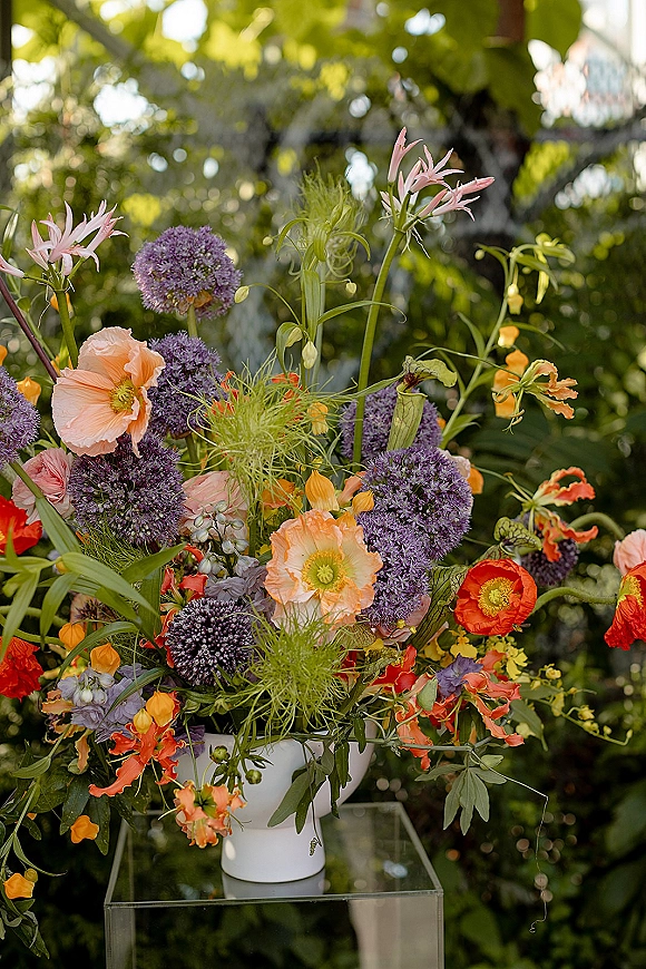 Wedding floral arrangement, tall wedding centerpiece with poppy and allium blooms in orange and purple on a clear pedestal in a sunlit garden setting