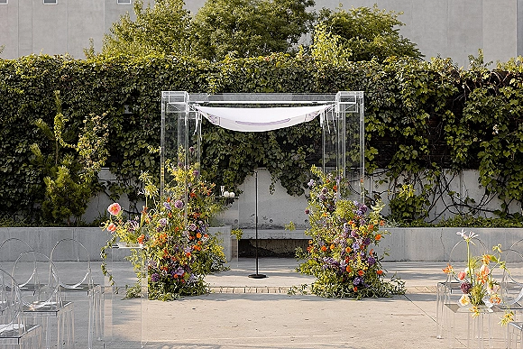 Ceremony altar with clear acrylic chuppah draped in white fabric, colorful florals and greenery, set before an ivy wall backdrop