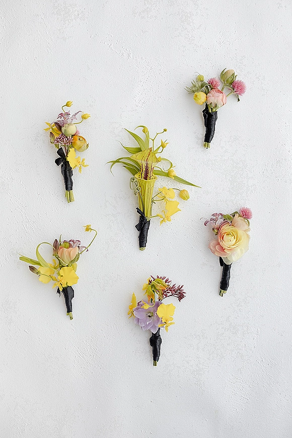 Wedding boutonnieres arranged on a white textured wall, featuring roses, orchids and ranunculus with greenery and black ribbon wrap