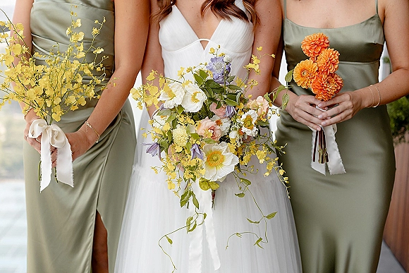 Bride and bridesmaids in sage green dresses holding colorful wildflower bouquets with ribbon wraps, standing amid outdoor greenery
