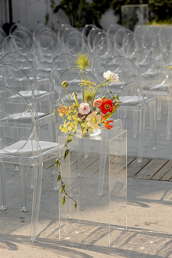 Ceremony aisle decor with an acrylic aisle pedestal holding a colorful wildflower arrangement in a glass bowl beside clear chairs on an outdoor patio