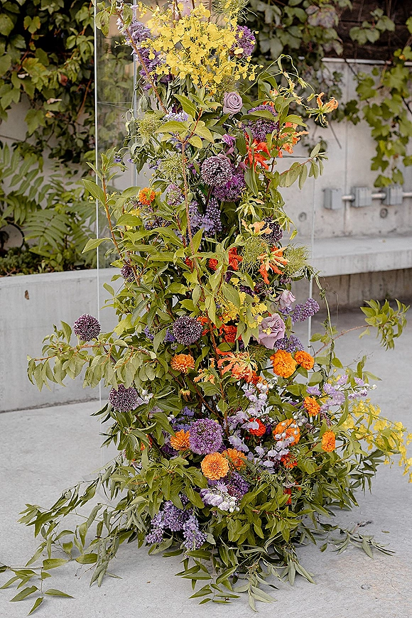 Wedding floral installation with hanging flowers and greenery on a clear acrylic frame against a concrete wall with climbing vines