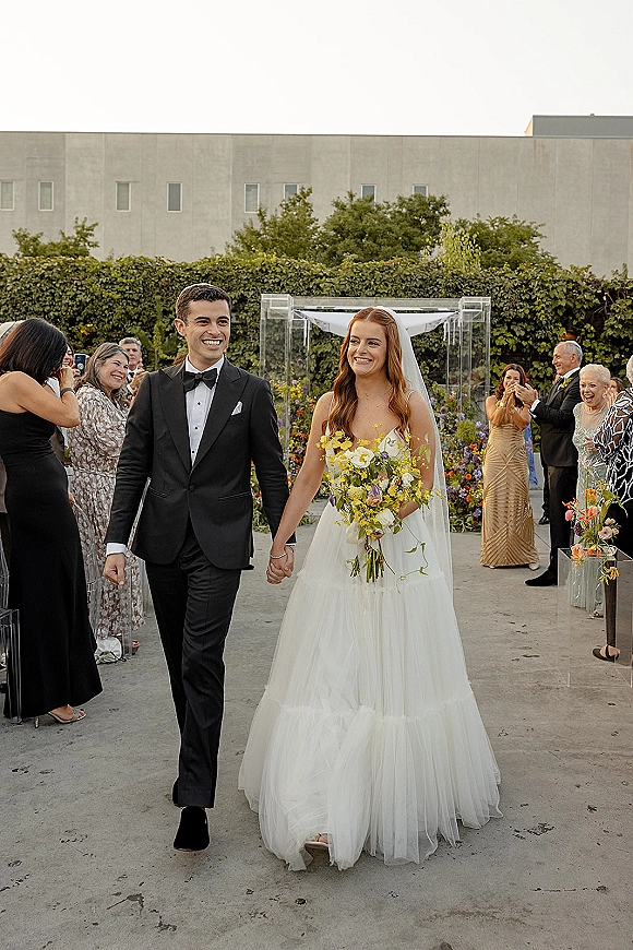 Wedding recessional as bride and groom walking aisle holding hands, her yellow wildflower bouquet and long veil in an outdoor courtyard with guests cheering