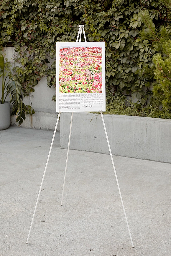 Wedding welcome sign on an easel with watercolor artwork and printed text, framed in white against an ivy-covered concrete wall backdrop