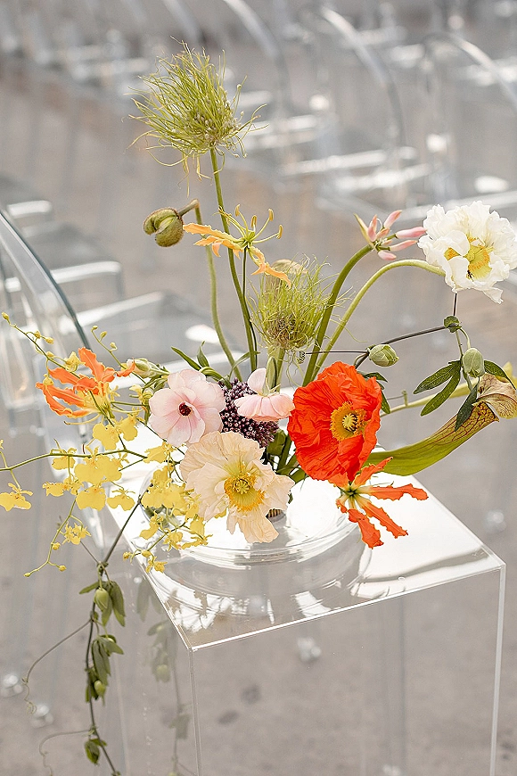 Wedding floral arrangement with ceremony aisle flowers in a glass vase on a clear acrylic pedestal, wildflowers in sunlight by clear chairs