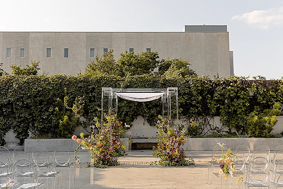 Ceremony setup with an acrylic chuppah draped in fabric, colorful florals and greenery garlands, set before an ivy-covered wall outdoors