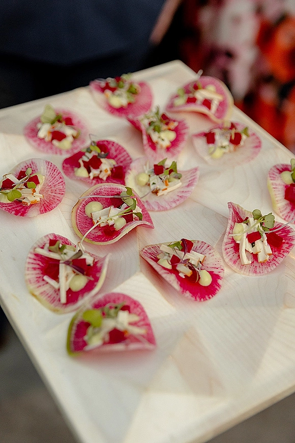 Wedding appetizers arranged as passed hors d'oeuvres on a wooden serving board with radish slices and microgreens against dark fabric backdrop