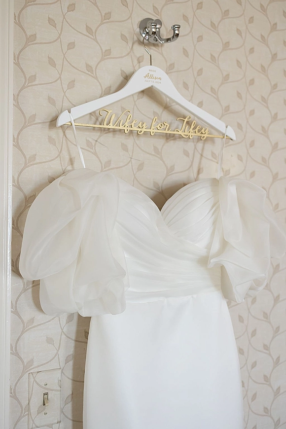 Wedding dress hanging from a gold wire hanger on a wall hook against patterned wallpaper, highlighting the clean white bridal gown silhouette