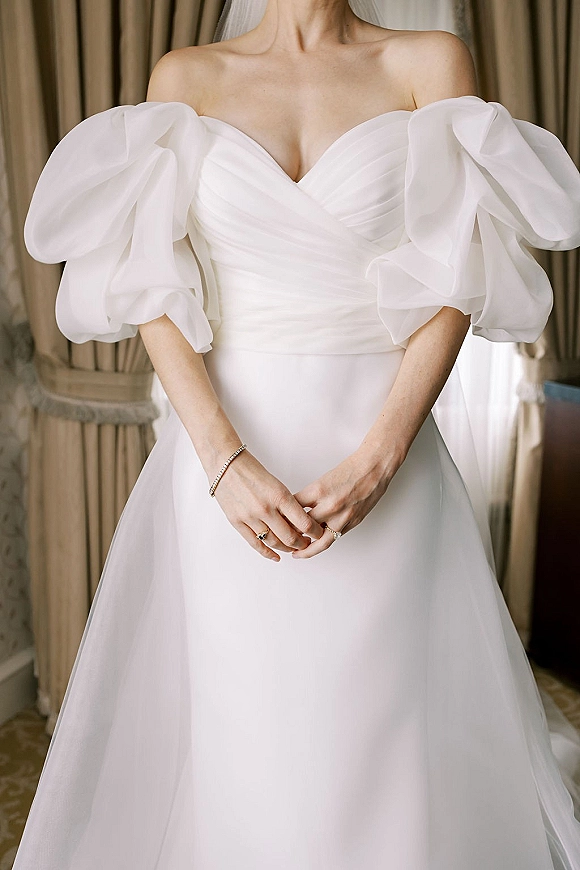 Wedding dress with off the shoulder puff sleeves, pleated bodice and sweetheart neckline, shown with bracelet and rings by draped curtains indoors