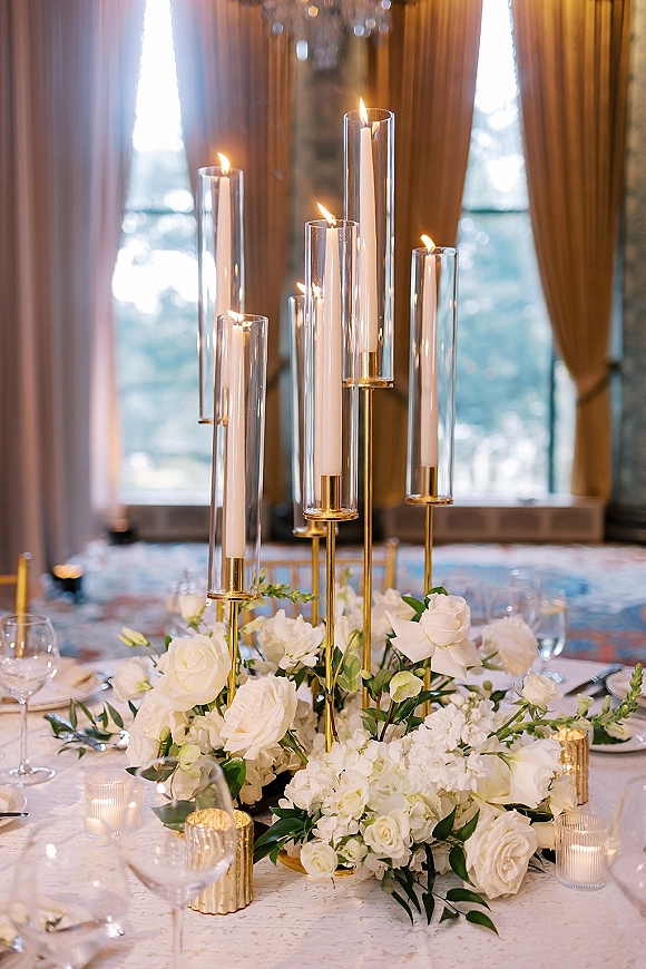 Reception centerpiece with tall candle centerpiece of taper candles in glass hurricanes, white roses and greenery on a chandelier-lit table