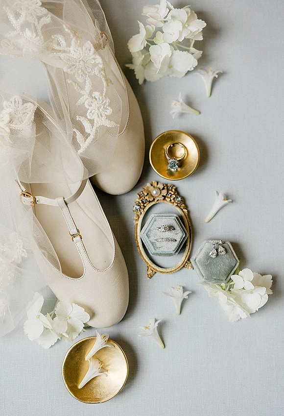 Wedding details flat lay with bridal accessories arranged on gray fabric, featuring ivory shoes, lace veil, rings in boxes, and white flowers