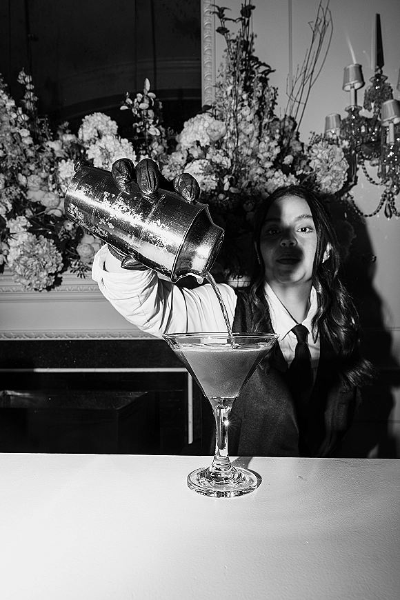 Wedding cocktail moment with a gloved bartender shaking a martini at a wedding bar setup beneath a chandelier and large floral display indoors