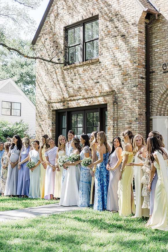 Bridesmaid group photo showing a wedding party lineup in mismatched dresses holding small white bouquets outside a brick house on a lawn