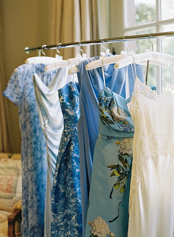 Bridesmaid dresses in mixed blue tones hang on a rack beside a lace-trim wedding dress, lit by soft daylight near a window